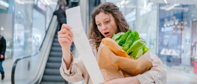 A woman holding groceries and looking shocked at the bill.