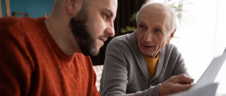 Senior father with adult son on sofa at home, reading and discussing documents.
